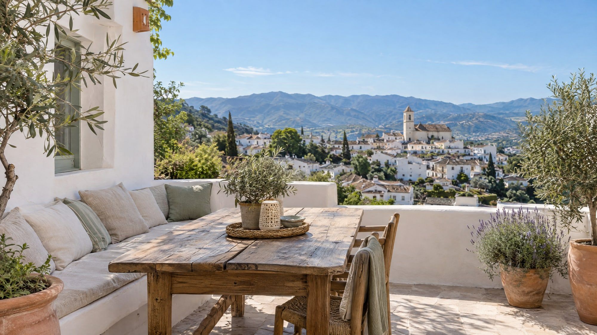Terraza encalada con mesa de madera, olivos en maceta y vistas a un pueblo